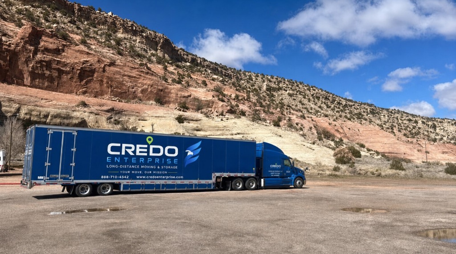 Credo Enterprise long-distance moving semi-truck with navy trailer branding parked in a desert landscape under a blue sky
